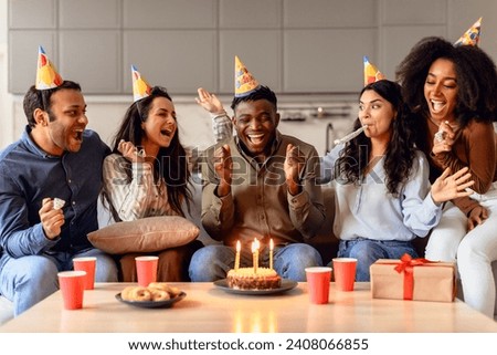 Happy multiracial students celebrating birthday of black guy, cheering while he makes wish and blowing candles on cake in modern kitchen interior. Bday party with true friends