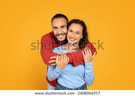 Attractive millennial couple hugging on orange studio background, smiling at camera. Happy young man and woman embracing, posing together. Marriage concept