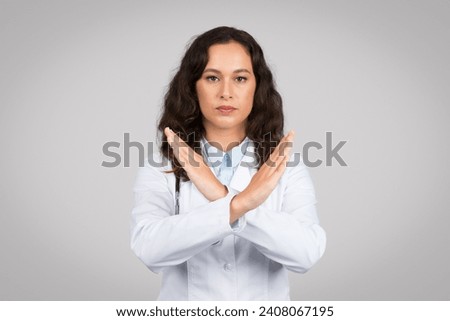 Serious female doctor in white coat making X sign with her arms, signaling denial or prohibition, wearing white lab coat with stethoscope, grey background Royalty-Free Stock Photo #2408067195