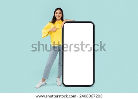 Cool cheerful stylish pretty young brunette woman pointing at huge phone with white blank mockup screen, showing nice online offer or recommending mobile app, blue background, copy space