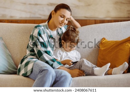 Mom and child relaxing with smartphone on cozy couch, mother and preschool daughter sharing a quiet moment together on comfortable sofa flanked by colorful cushions, watching cartoons together