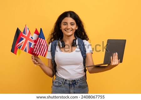 Happy young indian woman student with many different flags holding computer, isolated on yellow background, studio. Online school, course, education, ad, offer, modern technology