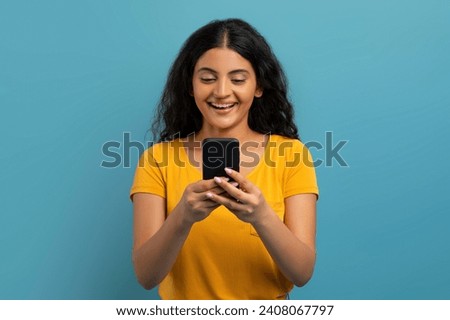Positive pretty curly young indian woman wearing yellow t-shirt using cell phone isolated on blue studio background, texting her friends, scrolling, websurfing, using newest mobile app Royalty-Free Stock Photo #2408067797