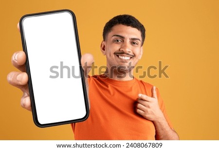 Cheerful latin man showing big cell phone with white blank screen and pointing at it, recommending online offer, mobile app, standing on yellow studio background. Check this deal concept