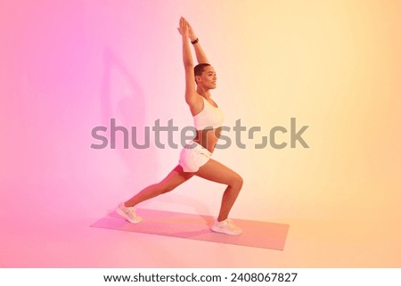 A fit latin woman with a shaved head practices a yoga warrior pose with precision and grace on a mat, wearing a white sports bra and shorts against a pink and yellow gradient Royalty-Free Stock Photo #2408067827