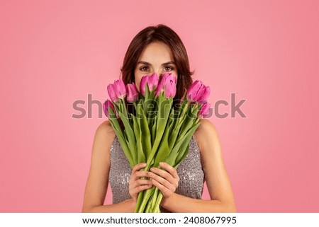 Enigmatic woman peering over a lush bouquet of pink tulips, her eyes captivating with a hint of mystery against a soft pink background. Spring holiday celebration event, ad and offer Royalty-Free Stock Photo #2408067995