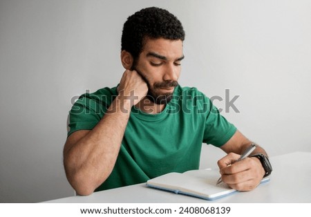Serious man with a beard in a green t-shirt writing in a notebook, resting his chin on his hand, looking focused at his work, at a white desk with a smartwatch on his wrist