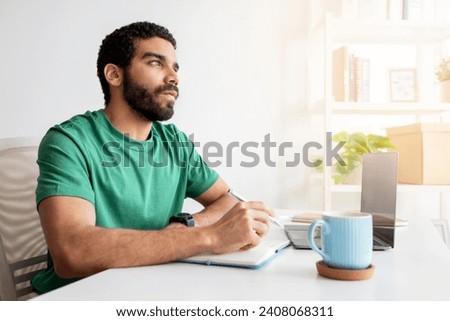 Thoughtful middle eastern man with a beard and green t-shirt looking away while writing in a notebook at his workplace, with a laptop and a mug on the desk in a sunny home office