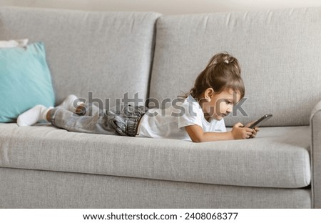 Cute toddler girl using smartphone while lying on comfortable sofa at home, little female kid playing games or watching cartoons, showcasing early engagement with technology in modern digital age