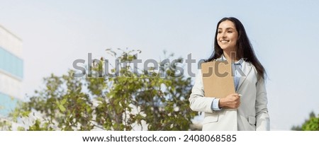 Professional glad caucasian young woman outdoors holding a clipboard, dressed smartly in a suit jacket, looking optimistically towards her future in the business world, panorama Royalty-Free Stock Photo #2408068585