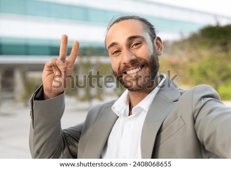 Friendly young businessman in suit making peace sign while taking selfie outdoors, happy male entrepreneur displaying positivity and carefree attitude in professional setting, closeup shot