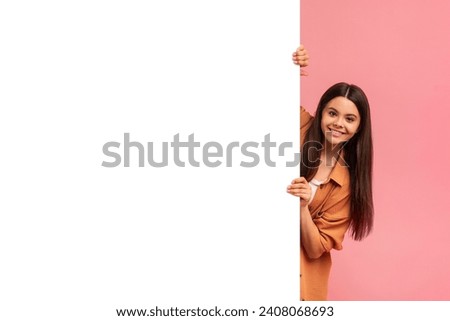 Friendly teenage girl peeking out from behind white advertisement board set against contrasting pink background, happy teen female creating playful and inviting image with copy space