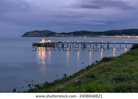 Long historic pleasure pier at dusk with lights reflecting in the water. Llandudno seaside resort, North Wales Royalty-Free Stock Photo #2408068821
