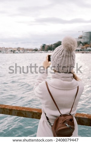 Girl with a backpack stands on the pier and takes pictures of the sea coast. Back view