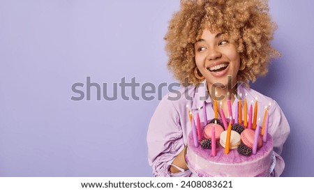 Studio shot of glad woman with curly bushy hair holds big tasty cake looks aside happily smiles broadly wears festive clothing celebrates birthday isolated over purple background copy space for text