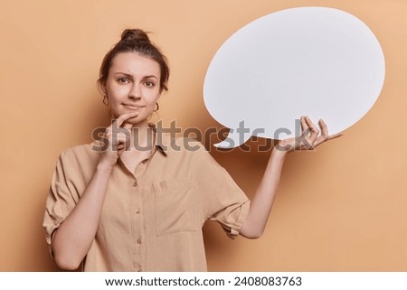 Thought quote and opinion concept. Pensive young woman with hair bun holding blank white speech bubble shows copy space for text or feedback on word cloud poses against brown studio background