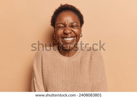 People and happiness. Indoor photo of young glad smiling broadly African american girl with closed eyes standing isolated in centre on beige background wearing casual sweater with short curly hair Royalty-Free Stock Photo #2408083801