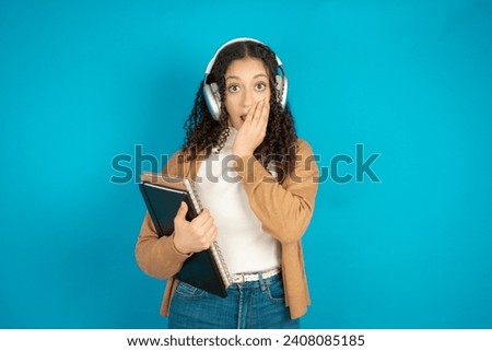 Shocked Beautiful student girl holding books wearing formal clothes stares fearful at camera keeps mouth widely opened wears wireless stereo headphones on ears