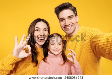 Close up young parents mom dad with child kid girl 7-8 years old wear pink sweater casual clothes do selfie shot mobile cell phone show ok okay isolated on plain yellow background. Family day concept