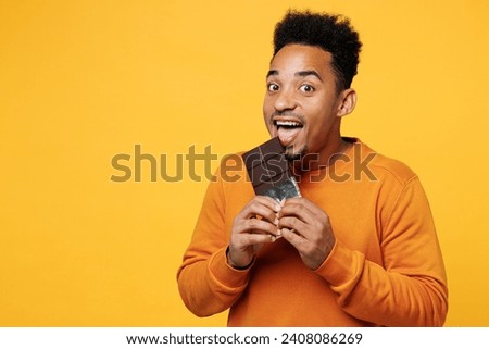Young happy man wear orange sweatshirt casual clothes hold eat biting bar of chocolate isolated on plain yellow background studio portrait. Proper nutrition healthy fast food unhealthy choice concept Royalty-Free Stock Photo #2408086269