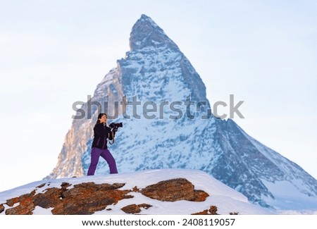 Traveler photographer woman with camera taking pictures of landscape of Swiss Alps on winter day, standing on snowy stone mountain plateau against sharp Matterhorn peak