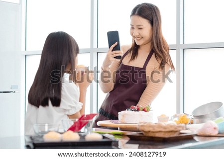 Asian beautiful female baker pastry chef mother using smartphone taking photo of little girl daughter wearing apron standing smiling holding cupcake posing after finish decorating homemade tasty cake.