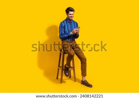 Full size photo of positive smart guy dressed striped shirt sit on chair read email on smartphone isolated on yellow color background
