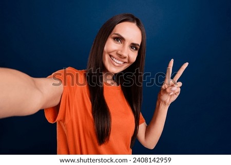 Photo of adorable excited woman dressed orange t-shirt smiling tacking selfie showing v-sign isolated dark blue color background
