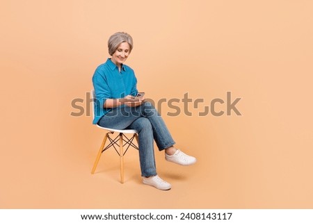 Full size photo of good mood smart woman dressed stylish shirt sit on chair look at smartphone isolated on pastel color background
