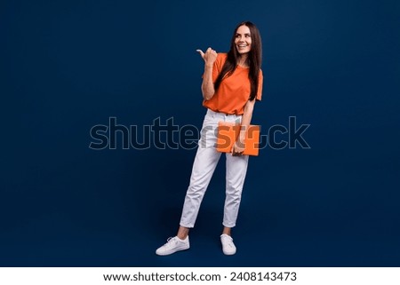 Full length photo of adorable dreamy woman dressed orange t-shirt holding gadget pointing empty space isolated dark blue color background