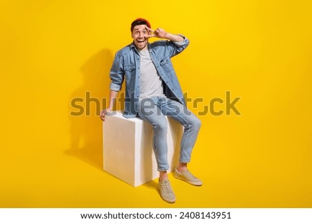 Full length photo of excited positive man dressed jeans shirt sitting white cube showing v-sign cover eye isolated yellow color background