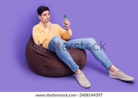 Full size photo of attractive young man sit bean bag unsure hold device dressed stylish yellow clothes isolated on purple color background