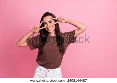Portrait of lovely satisfied girl toothy smile arms fingers demonstrate v-sign near eyes empty space isolated on pink color background