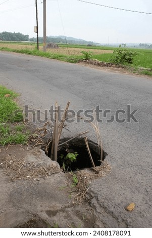 East Java, Indonesia. A deep open hole on the edge of an asphalt road in a village near a rice field area, on either side of the road there are wild grasses