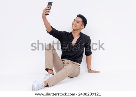 A young stylish asian man sitting relaxed on the floor, one knee up, taking a selfie with his phone. Candid studio shot isolated on a white background.