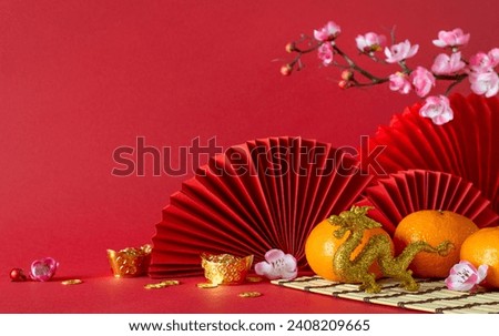 Traditional New Year tableau: side view feng shui elements, gold coins, dragon ornament, tangerines, sakura flowers, and fans arranged on red surface, providing a perfect canvas for text or promotions