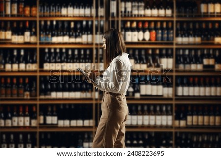 Female expert sommelier locating in winery sphere analyses various varieties of red and white wine, located in wine cellar, making notes in tablet.  Royalty-Free Stock Photo #2408223695