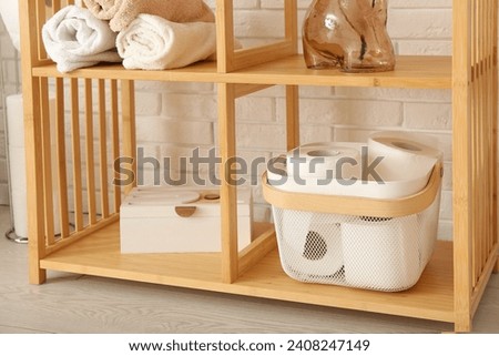 Wooden shelving unit with toilet paper rolls, clean towels and decor near light brick wall, closeup Royalty-Free Stock Photo #2408247149