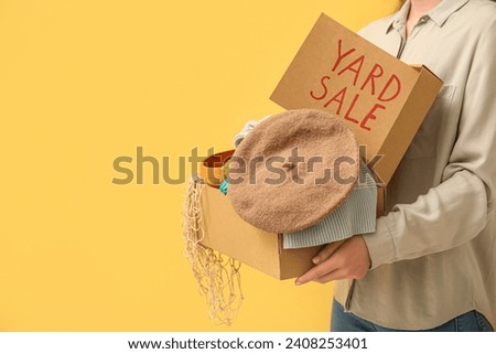 Woman holding box of unwanted stuff and cardboard sign with text YARD SALE on yellow background