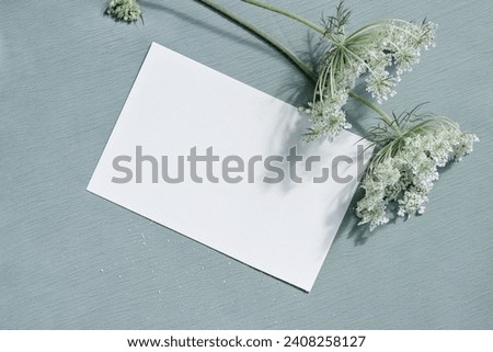 Beautiful white blooming flowers and blank paper card mockup on light blue textured cloth background. Aesthetic floral greeting postcard for Mothers day, Valentines day, wedding invitation template.