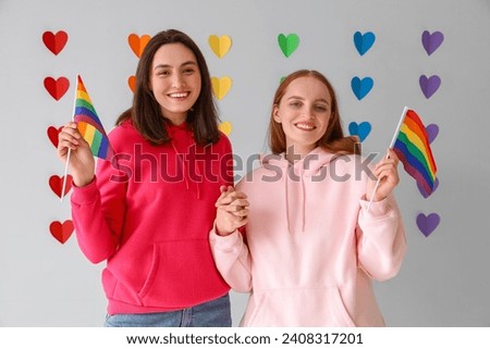 Young lesbian couple with LGBT flags against rainbow hearts on light background. Valentine's Day celebration
