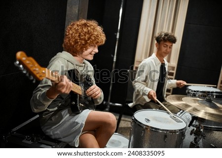 focus on cheerful red haired teenage boy playing guitar next to his blurred friend playing drums Royalty-Free Stock Photo #2408335095