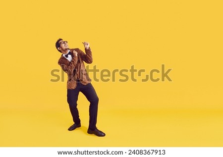 Happy funny man on yellow colour advertising text copyspace background. Full body studio shot of cheerful young ethnic guy in trendy leopard jacket with bowtie dancing and looking up at something Royalty-Free Stock Photo #2408367913