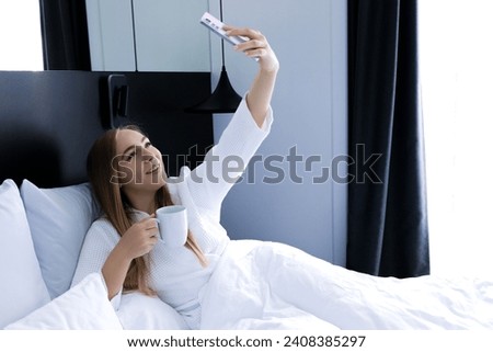 Young smiling girl in the morning lying in bed and taking selfie on her smartphone. Social media, lifestyle and technology concept.
