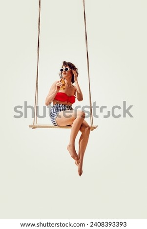 Beautiful, elegant young brunette woman in stylish vintage swimsuit, sunglasses sitting on swing with lemonade against white background. Concept of summer vacation, travelling, retro style, fashion