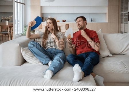 Suffering from heat family couple sitting on couch waving paper fans at home. Tired exhausted man woman feel bad in hot summer weather in room without air conditioner. Overheating, sultriness concept. Royalty-Free Stock Photo #2408398527
