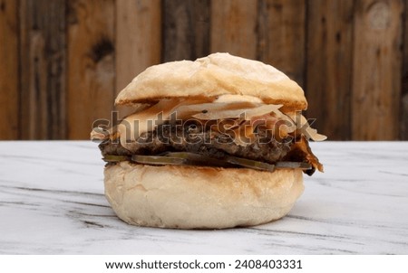 American food. Hamburgers. Closeup view of a burger with layers of meat, cheddar cheese, crispy bacon, onion, bittersweet cucumbers and sauce, on the white marble table with a wooden background.  Royalty-Free Stock Photo #2408403331