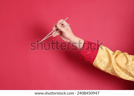 Happy Chinese new year. Asian Chinese energetic senior man wearing golden traditional cheongsam qipao or changshan dress with gesture of hand holding silver chopsticks isolated on red background. Royalty-Free Stock Photo #2408450947