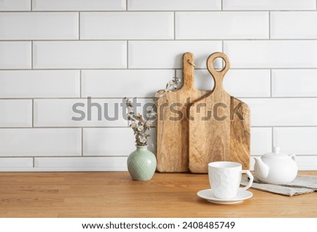 Wooden oak tabletop with a cup of tea and a teapot against a white brick background with cutting boards and a vase with dry branches. Cozy breakfast concept in Scandinavian style. Copy space.