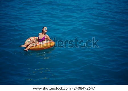A young beautiful girl in a bright swimsuit lies on a large inflatable ring and floats on the blue sea on a bright sunny summer day